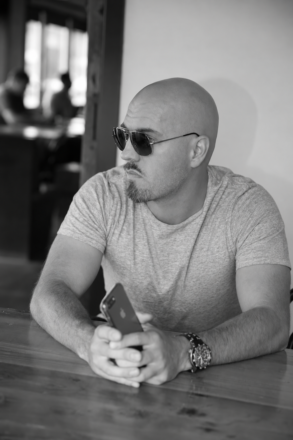 Liam Legault, founder of Liam Legault Design, seated at a wooden table holding a phone, wearing sunglasses and a plain grey T-shirt.